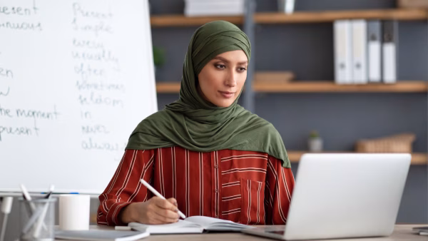 Islamic teacher in headscarf sitting at desk looking at laptop